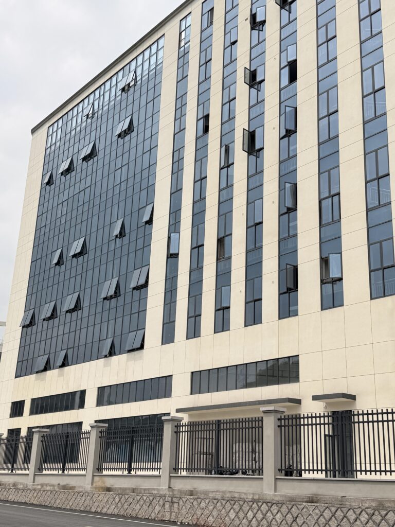 A multi-story beige industrial facility features rows of dark glass windows behind a black metal perimeter fence. This represents our high-capacity baby textile factory in China for OEM/ODM customized orders.