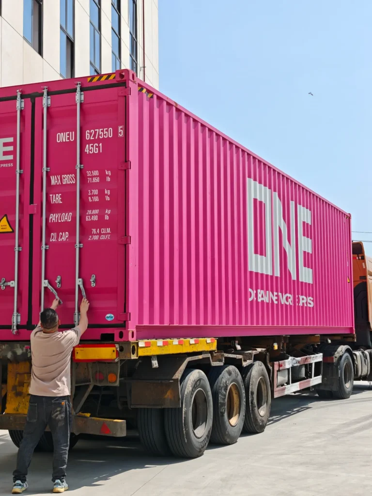 A worker fastens the locking bars of a magenta ONE shipping container on a flatbed trailer outside a factory building. As a Chinese baby textile factory, we provide full OEM/ODM customized export logistics.