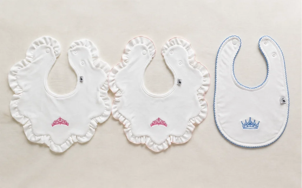 Our custom baby bib samples compare ruffled flower shapes, a rounded style, pink or blue crown embroidery, and varied edge finishes at our Hangzhou, China OEM/ODM factory with 40+ years of experience.