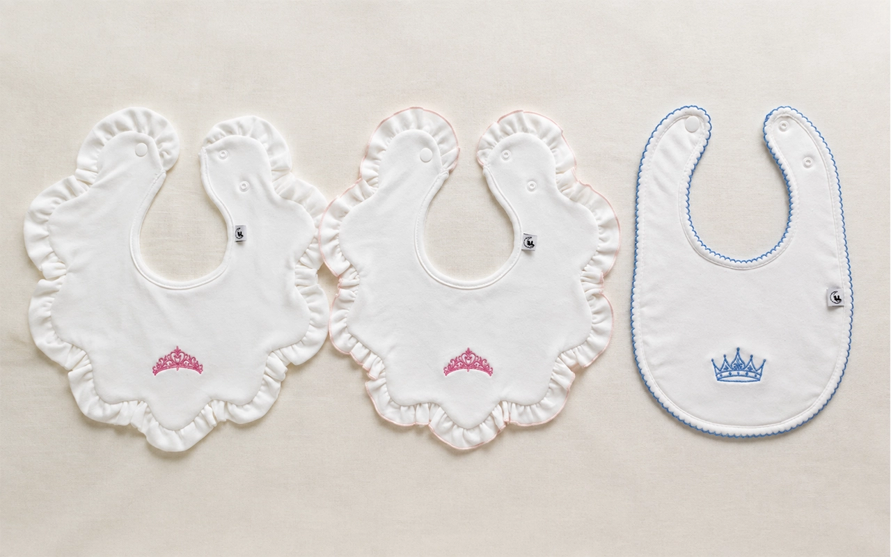 Our custom baby bib samples compare ruffled flower shapes, a rounded style, pink or blue crown embroidery, and varied edge finishes at our Hangzhou, China OEM/ODM factory with 40+ years of experience.