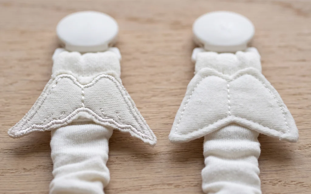 Our two ivory pacifier clips compare embroidered and quilted wing decorations above gathered fabric straps, showing how surface texture, stitching and wing scale change one coordinated product design.